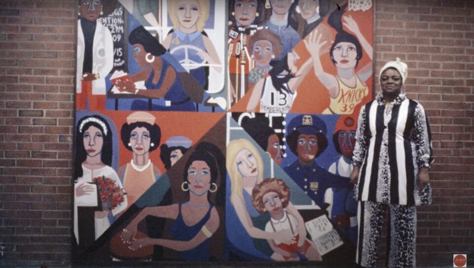 a colorful mural divided into eight parts depicting women in various professions. the artist wears a black and white outfit and stands next to the work 