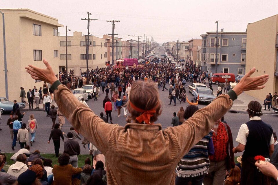 a man holds up his arms in a v with his back facing the camera, while people gather in the street in front of him