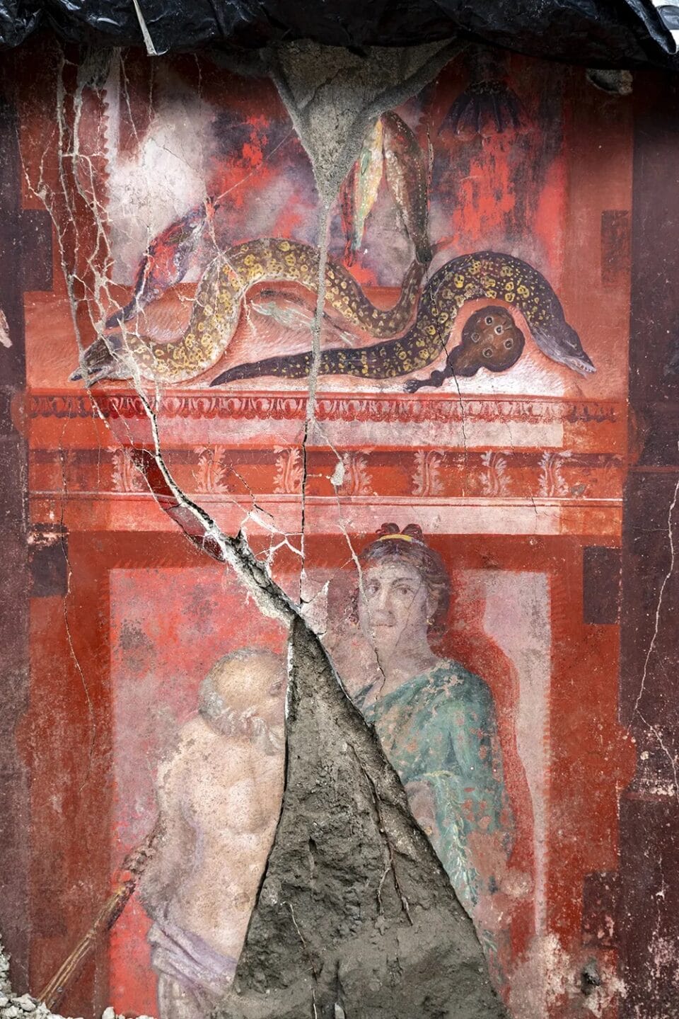a detail of a mostly-red fresco inside of an ancient banquet hall excavated in Pompeii, featuring a woman with an architectural detail above her, on which sit a couple of snakes