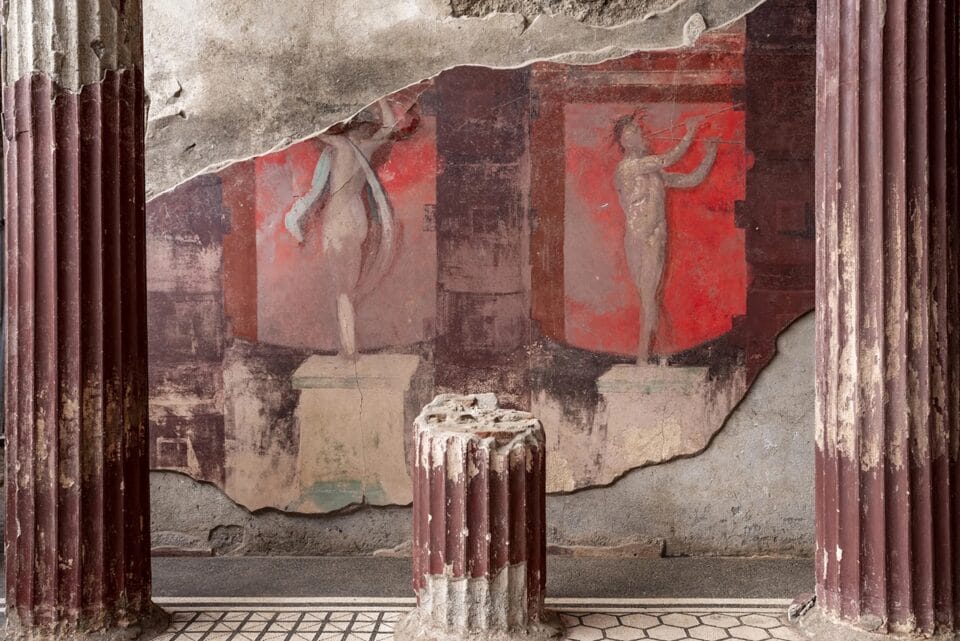 a detail of a mostly-red fresco inside of an ancient banquet hall excavated in Pompeii, featuring figures in painted architectural niches, foregrounded by large painted columns