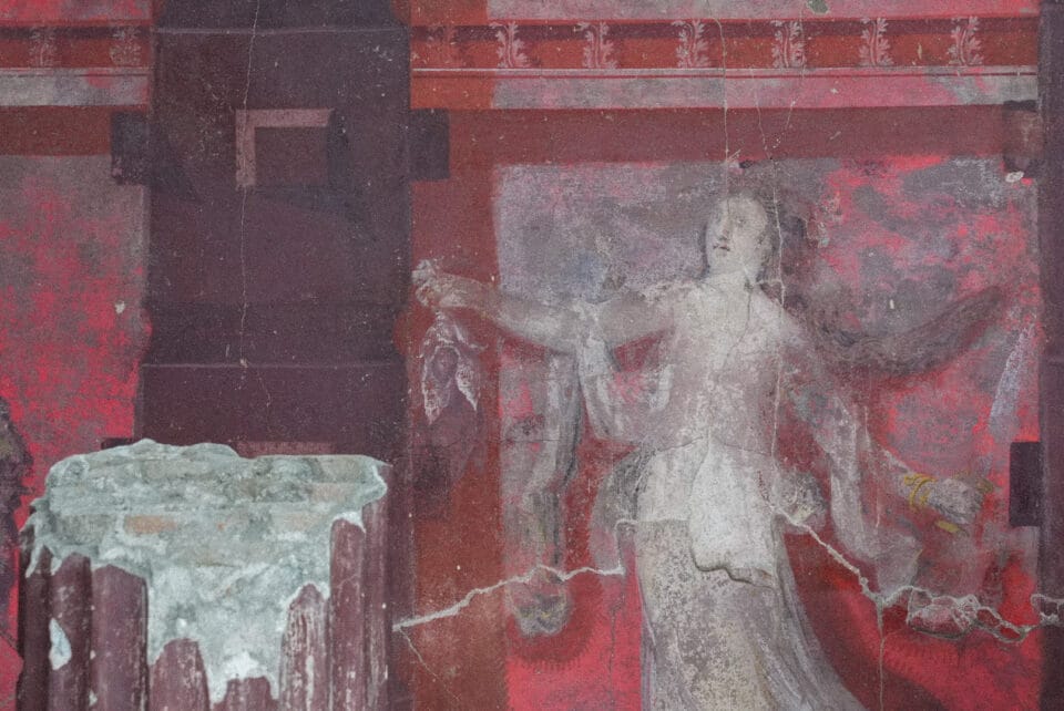 a detail of a mostly-red fresco inside of an ancient banquet hall excavated in Pompeii, featuring a female figure in a gown inside of a painted architectural niche, foregrounded by a large painted column