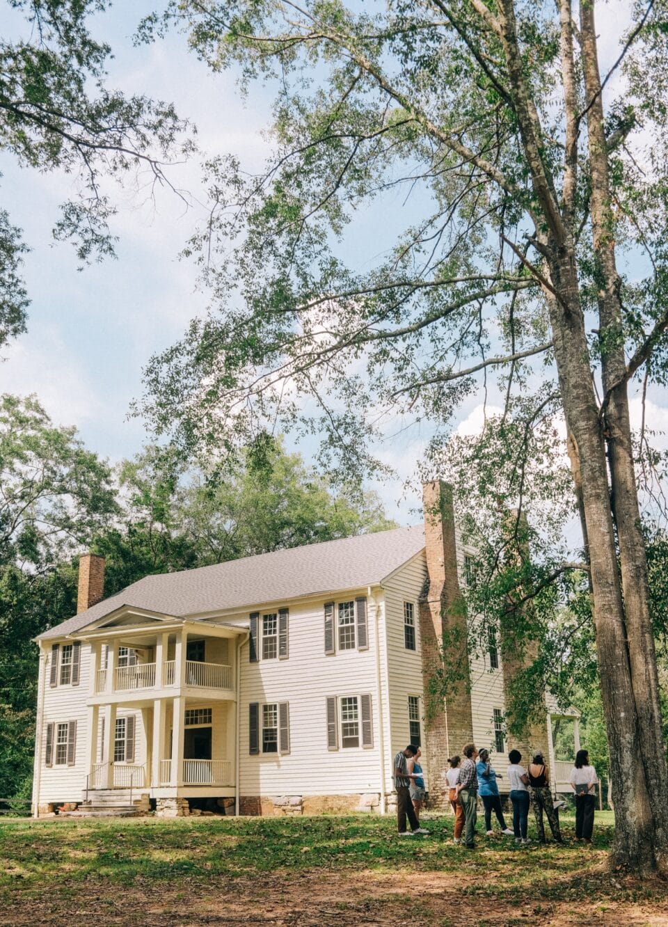 people gather under a tree outside an antebellum mansion