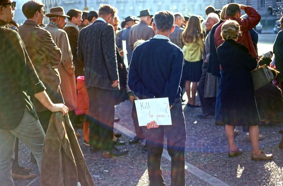 a man holds a sign behind his back at a rally that says "kill for peace?"
